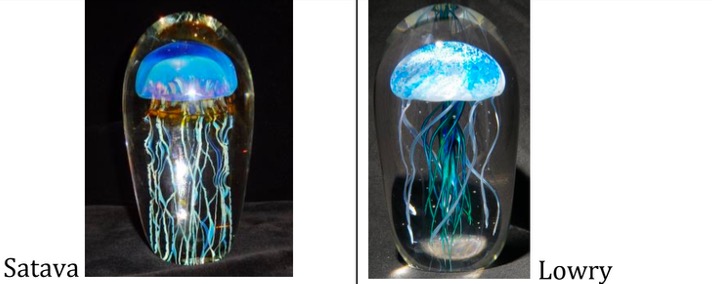 Two glass sculptures of jellyfish. Left, labeled “Satava,” features a domed blue-and-purple jellyfish with long, thin, wavy tentacles suspended in clear glass. Right, labeled “Lowry,” shows a blue-and-white jellyfish with more numerous, flowing tentacles in clear glass. Both pieces appear illuminated against a dark background.